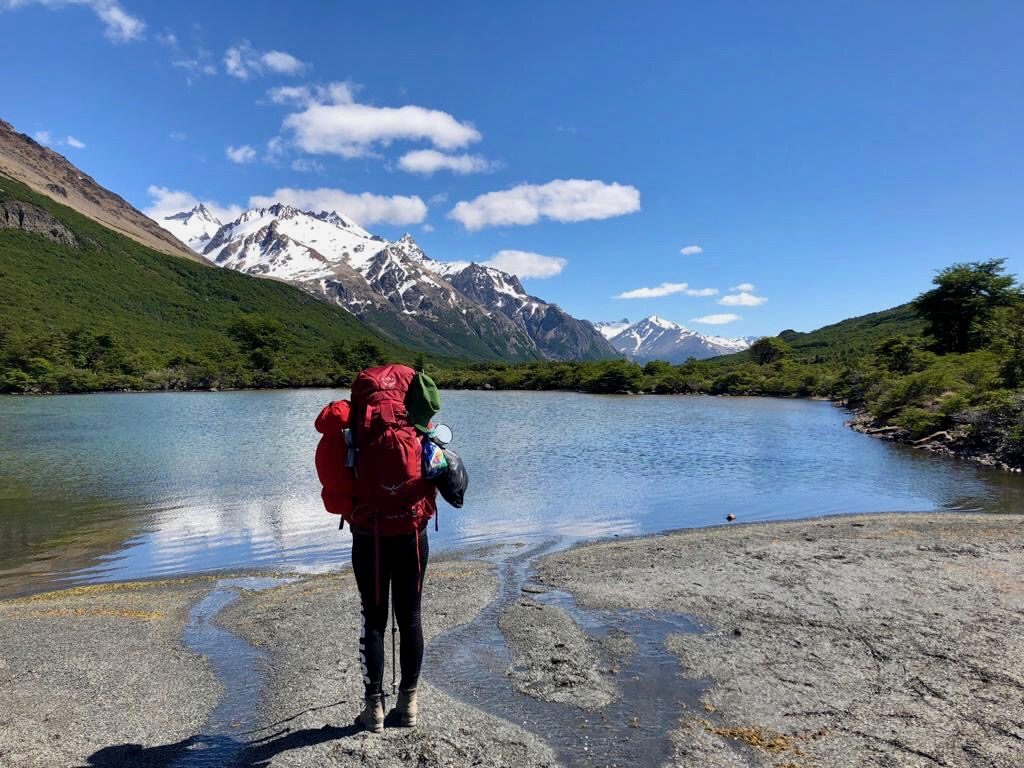 Charlotte vom Blog Rausgier steht mit einem großen, roten Trekkingrucksack an einem See vor Gebirgspanorama in Patagonien. 