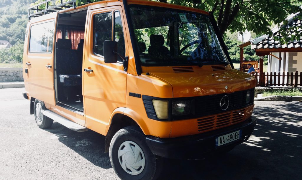 Ein orange-gelber Mercedes-Bus vor grünem Berg und Häuschen bringt Trekker zum Peaks of the Balkans.
